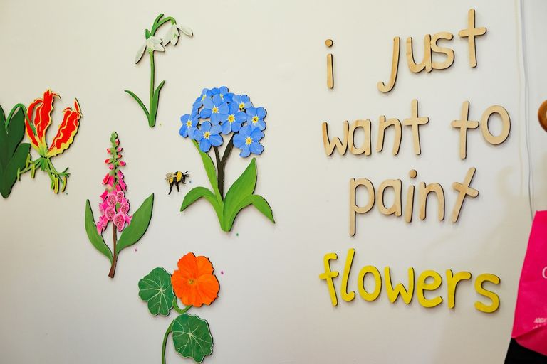 Art exhibition wall displaying colorful painted cutout flowers alongside wooden letters spelling "i just want to paint flowers."